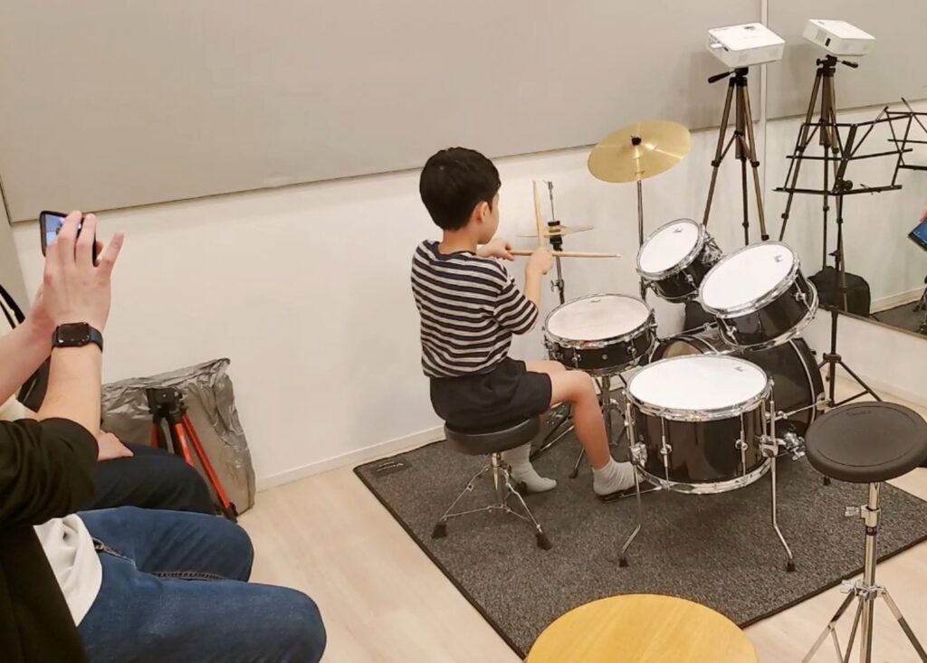小学生が先生のマネをしてドラムを叩こうとしているシーン